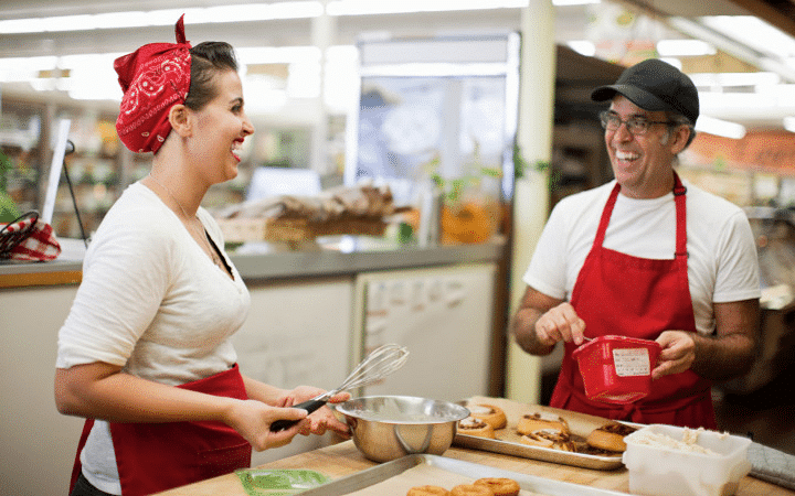 A woman and a man are wearing red aprons while they work together to ice freshly made donuts and cinnamon rolls in the kitchen of a small business.