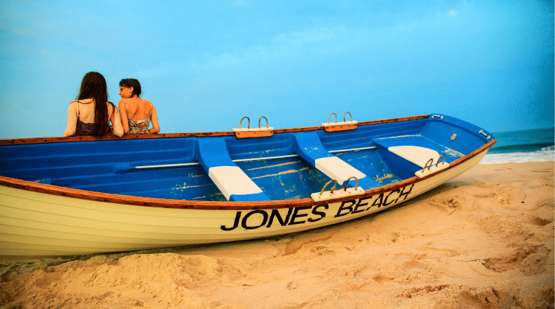 Two girls are chatting while they lean against a white and blue wooden row boat on Jones Beach in Long Island, New York. The words “JONES BEACH” are painted on the side of the boat in black letters. 