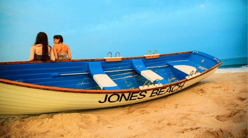 Two girls are chatting while they lean against a white and blue wooden row boat on Jones Beach in Long Island, New York. The words “JONES BEACH” are painted on the side of the boat in black letters.
