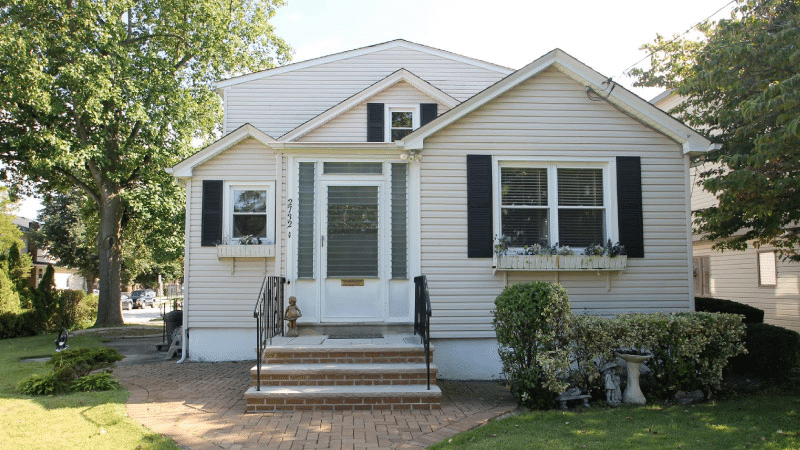 A cute, one-story residential home in Bellmore, New York, on Long Island. The house is white with black shutters, and there are three brick stairs leading up to the front door. A brick path surrounds the left side of the house and leads straight out from the entrance through the front yard.