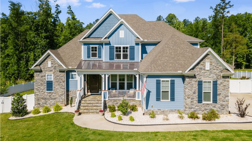 A new construction,  two-story home in Wake Forest, North Carolina. The exterior of the home is a combination of blue siding and tiles and natural stone veneer. The shutters are painted a darker blue and there’s a curved path leading up to the entrance stairs. The home backs up to a fenced yard and a lush forest.