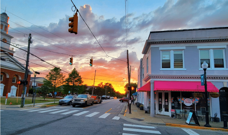 A street corner in downtown Apex, North Carolina, at sunset. Cars are stopped at an intersection as the sun is setting behind them, turning the sky a fiery orange and red. 