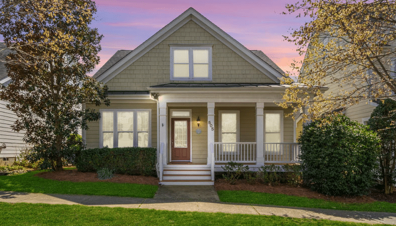 A quaint bungalow in Holly Springs, North Carolina. The home has wooden shingles on the side, painted a light cream. There’s a covered porch and a red door. The sun is setting behind the home, coloring the sky in pinks and blues. 