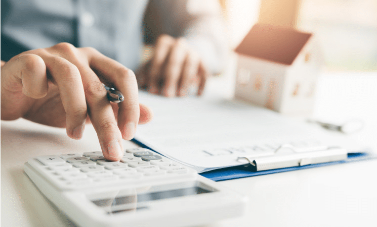 A closeup of a man using a calculator to calculate his home-buying budget. He is holding a pen and there is a clipboard with papers and a small model of a house on the table.