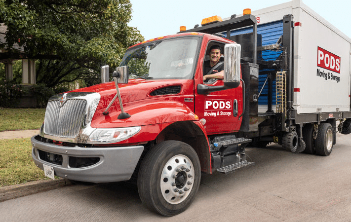 Two men are carrying a loveseat into the back of a moving truck rental. There are a couple of moving boxes in the foreground of the image.