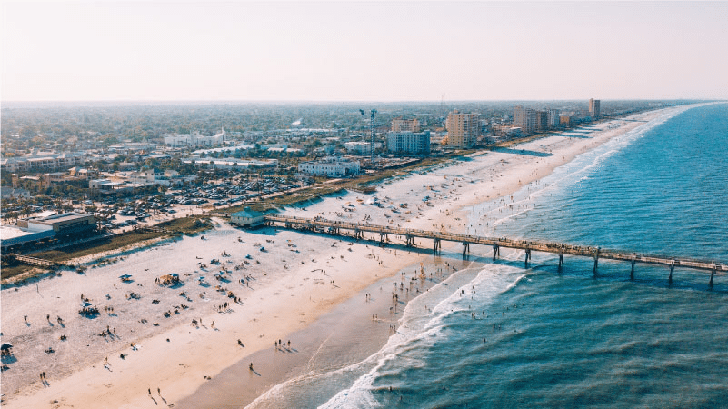 Aerial view of Jacksonville Beach, Florida, on a beautiful summer day. People are sunbathing, swimming, and walking along the shore and the pier. 