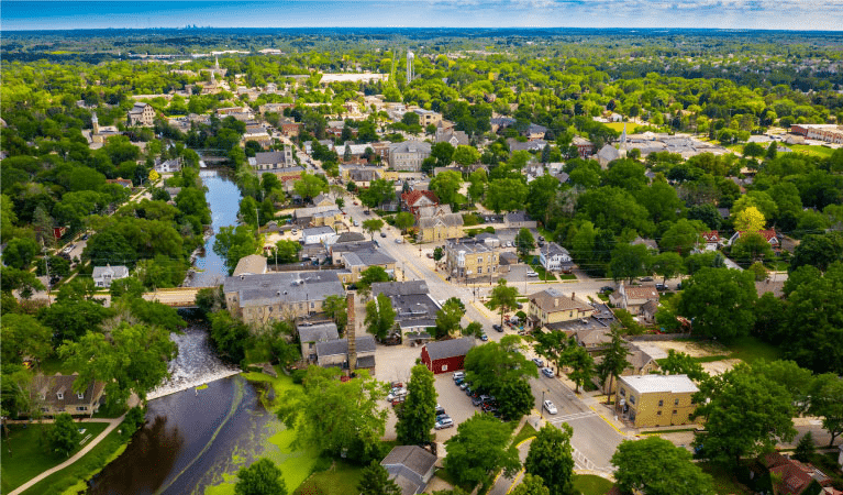 Aerial view of Cedarburg, Wisconsin, on a summer day. The sky is a lovely blue, the trees are bushy and green, and there’s a small waterway running through the town.