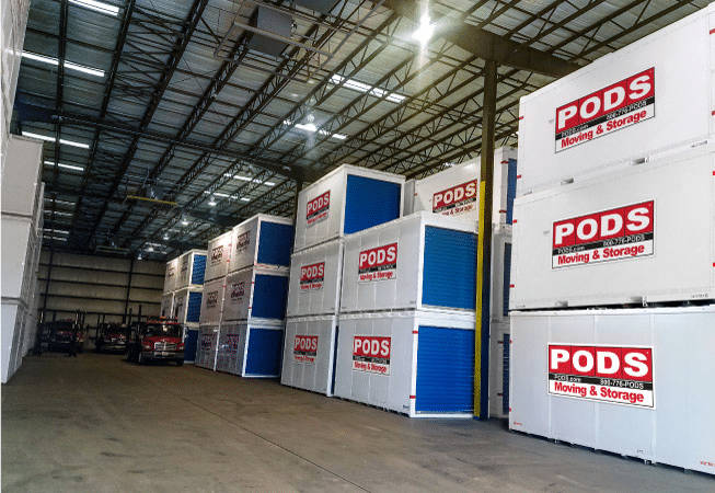 View of the inside of a PODS Storage Center in Greenville, SC. PODS containers are neatly stacked along the aisles and a few trucks are parked in the back of the storage center.