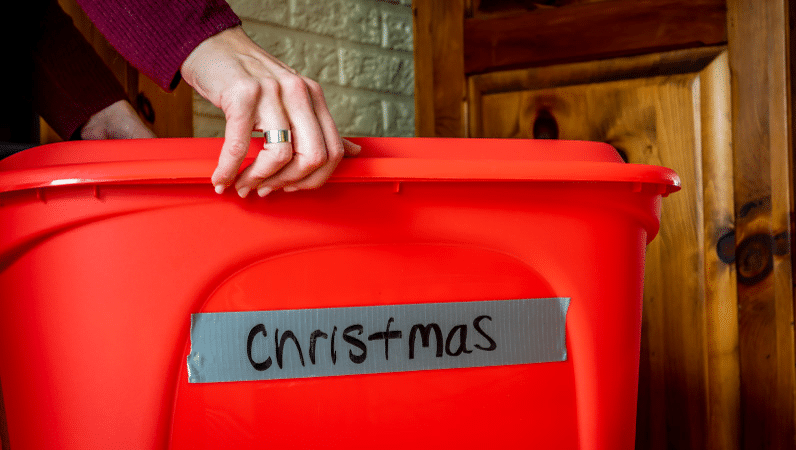 Close-up view of a pair of hands carrying a red plastic bin. There’s a piece of duct tape across the side of the bin labeled, “Christmas.” 