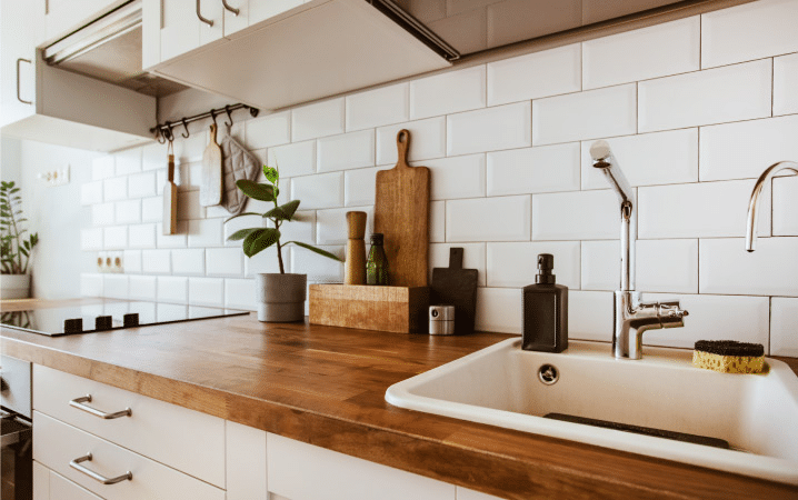 A modern farmhouse kitchen with a butcher block counter in a rich natural color. The cabinets, sink, and backsplash are all white, contrasting nicely with the rich wood color of the counter. 