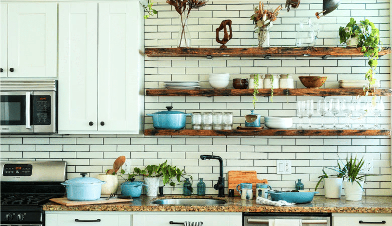 A modern farmhouse kitchen with floating wood shelves, white cabinets, and a white subway tile backsplash. There are house plants, pots, and dishes on the counter and on the shelves. 