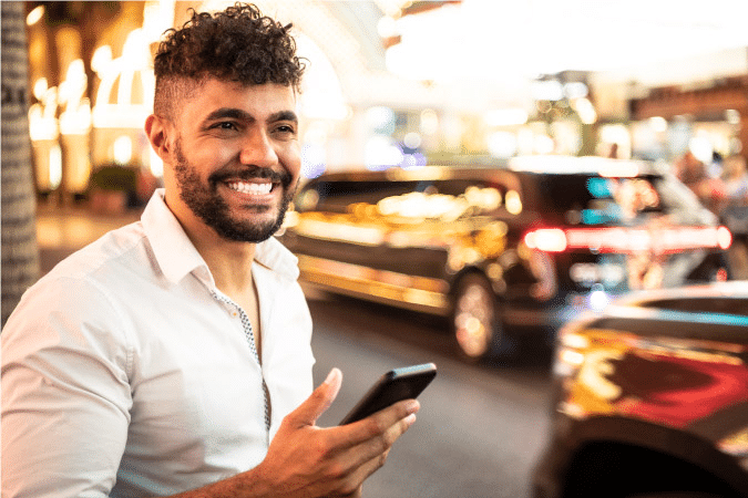 A young man with a chic haircut and a beard is holding his cell phone and smiling widely. The bright lights of the Las Vegas city streets are blurred behind him.