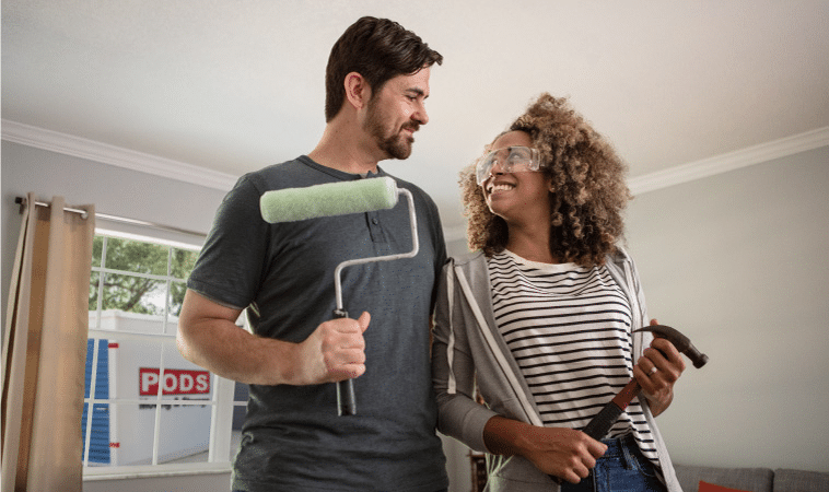 A man and woman are standing in their living room, smiling at each other. The woman is wearing protective eyewear and holding a hammer and the man is holding a paint roller. Through the window behind them, you can see a PODS portable storage container outside in the driveway. They’re preparing to do some work on their kitchen and will be using the PODS portable container to store things out of the way during the remodeling.