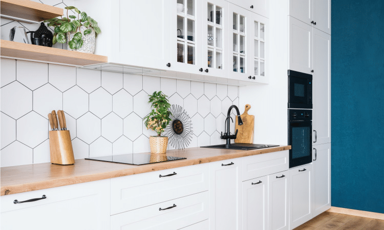 A modern farmhouse kitchen with white shaker-style and glass-front cabinets with metal handles. There’s a butcher block counter and the backsplash is made from white hexagonal tiles.
