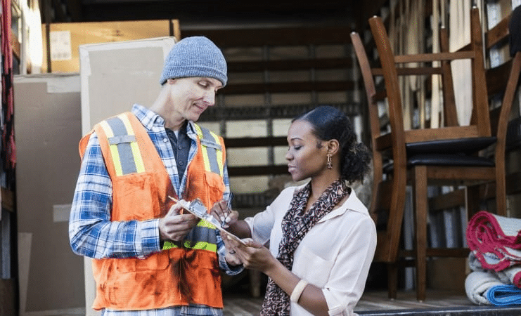 A woman signs paperwork as a professional mover holds the clipboard. They’re standing in front of a moving truck that is partially loaded with the woman’s furniture and moving boxes. 