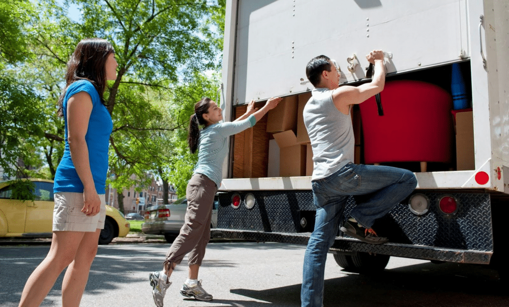 A man and a woman work together to open the rear door of a moving truck rental as another woman looks on. The door is about a third of the way open and a red chair and various moving boxes are visible inside the truck. 