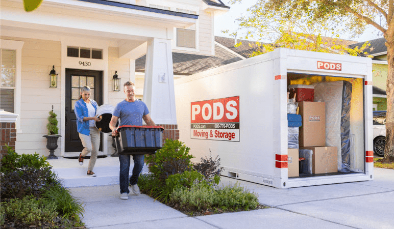 A man and woman are carrying a piece of furniture and a plastic tub out to their PODS portable moving container in the driveway. It’s almost fully loaded and you can see a mattress, cooler, and several moving boxes that are already packed in the container.