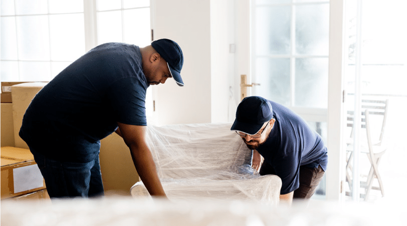 Two professional movers are lifting a piece of furniture secured in a protective wrap. They are in the middle of moving furniture and boxes out to a truck to be transported cross-country.