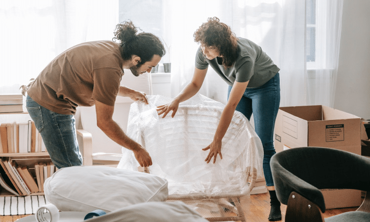 Two friends are working together to wrap a piece of furniture for a move. There is a bookshelf behind them and an empty moving box to the side.