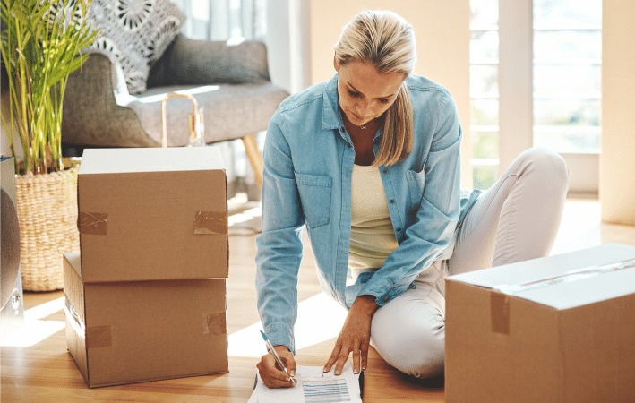 A woman is sitting in her living room beside some packed moving boxes. She is checking items off a list for her DIY move.