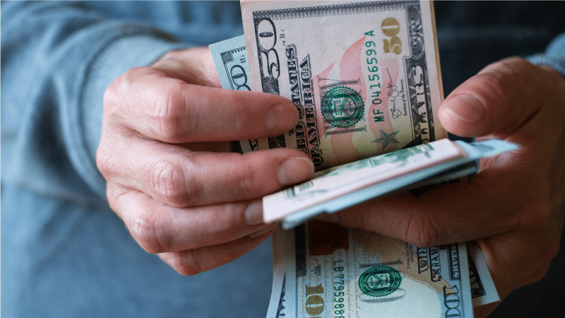 Close-up view of a man counting large U.S. bills. He’s holding a few hundreds and fifties and he’s wearing a gray, long-sleeved shirt.