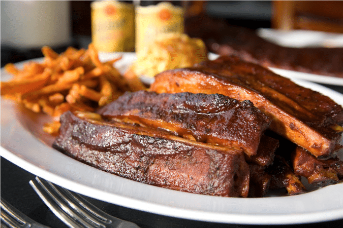 A close-up view of a plate of delicious-looking Kansas City barbecue ribs with seasoned french fries and a couple of cold beers in the background.