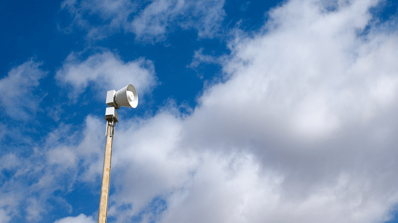 A white tornado siren is perched atop a tall, wooden pole. In the background is a bright blue sky with fluffy, white clouds.