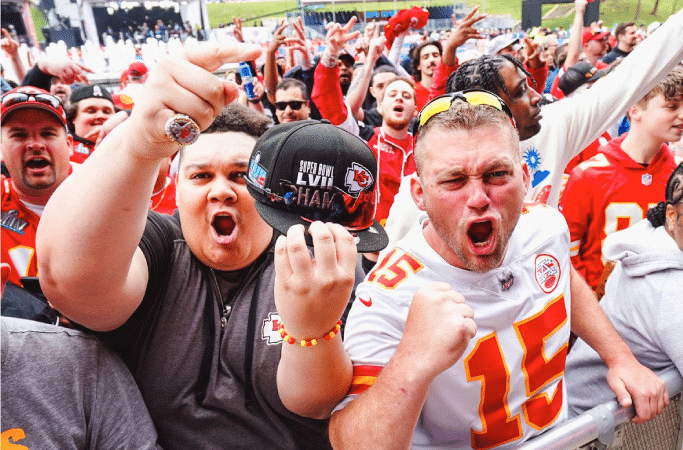 A crowd of excited Kansas City Chiefs fans cheer while posing for a picture. They’re decked out in team jerseys and colors.