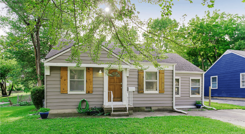 A small, single-family home in the Blue Hills neighborhood of Kansas City, Missouri. The exterior is made with gray siding and the door and window shutters are a natural wood. Sun is shining through the branches of a large tree in the front yard.