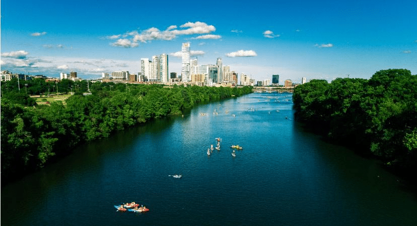 Locals are kayaking on Lady Bird Lake in Austin, Texas, with the city skyline in the distance. It’s a gorgeous summer day. The sky is a bright blue with occasional small puffs of white clouds. The trees along the river are lush with deep green leaves.