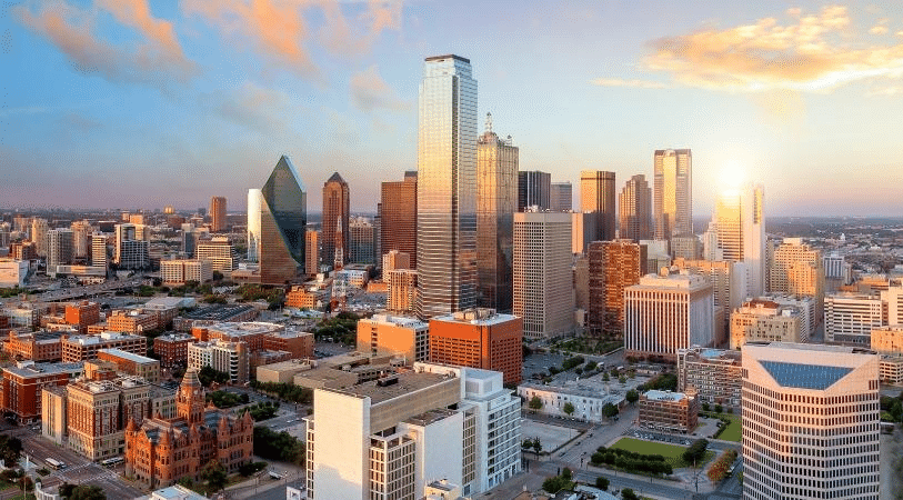 Aerial view of downtown Dallas during sunrise. Glass skyscrapers are reflecting the sky in a spectrum of colors from soft peaches to light blues.