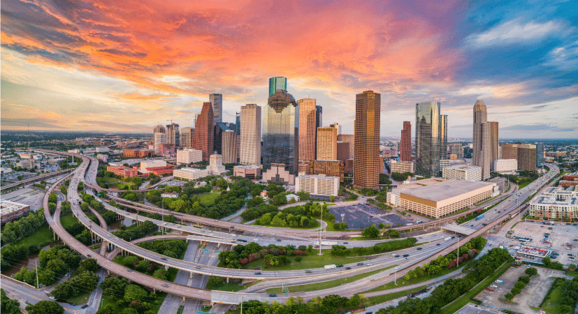 Aerial view of Houston, Texas, during sunrise. Freeways are wrapping around the downtown part of the city and mature trees are growing up in greenspaces between them. The clouds in the sky are reflecting a bright peach over the city but are blue in the distance.