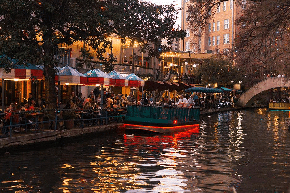 A small tour boat is floating down the San Antonio River as passersby dine and shop at the San Antonio River Walk.