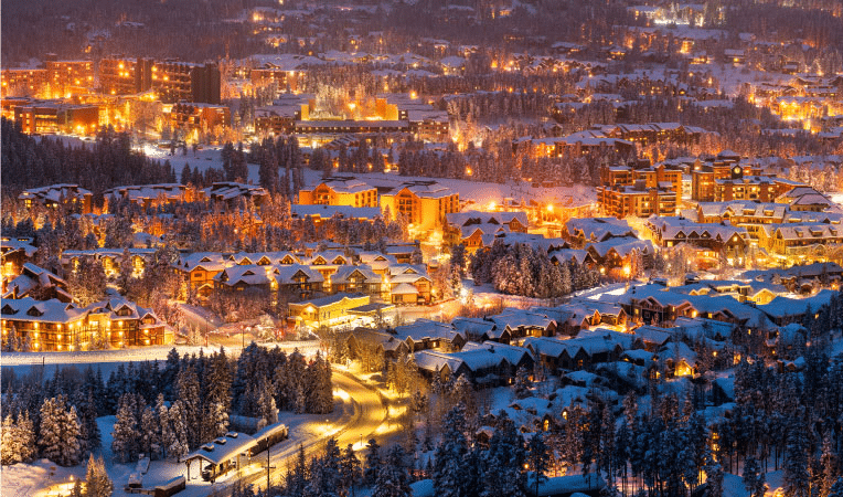 Breckenridge, Colorado, in the winter. Snow covers the trees and the tops of residential and resort buildings, and yellow lights in the windows illuminate the city. 
