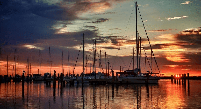 Sunset view of the Corpus Christi Marina. The sun is letting off a deep orange glow at the horizon, sending rays of light across the water and into the sky. Several sailboats are anchored with their masts reaching up into the sky. Dark clouds are moving in from the south.