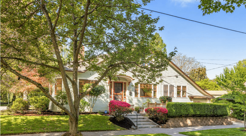 A one-story home in the Laurelhurst neighborhood of Portland, Oregon. The exterior is made of white siding and the windows and doors are framed with a bright, natural wood. The lawn is well kept and the shade of nearby trees is being cast on the house.