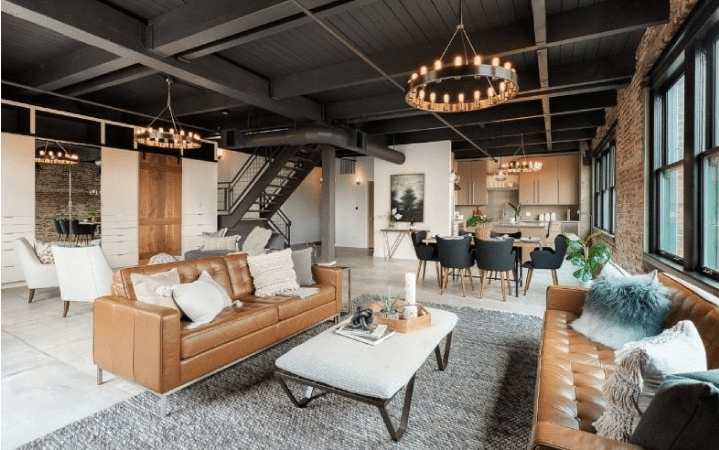 The chic interior of a condo at Chown Pella, a historic warehouse-turned-condominium building in the Pearl District. The living room is decked out in brown leather couches and a modern coffee table. There are medieval-style lighting fixtures throughout the living space, and the kitchen is visible in the background. Large windows are letting in natural light.