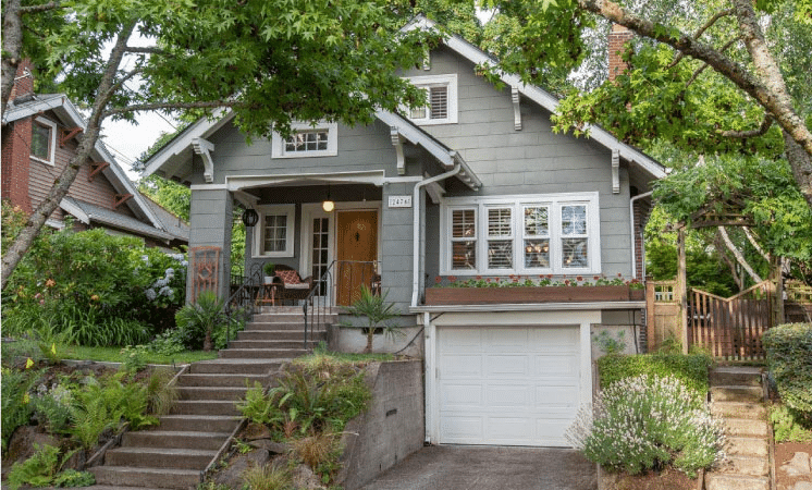 A two-story bungalow in the Hollywood neighborhood of Portland, Oregon. The home is built on a hill with a lush yard to the left of the home’s entrance. There are several steps leading up to the covered porch. On the first level, the garage is accessible from the street. The exterior of the home is made of gray wood siding and there is a long box of blooming spring flowers above the garage.