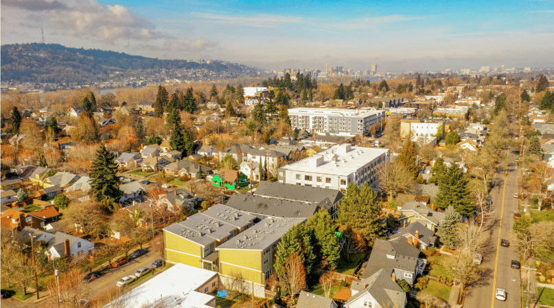 Aerial view of the Sellwood - Moreland neighborhood in Portland, Oregon, in the fall. It’s a bright day and the sun is reflecting off the residential and commercial buildings in the neighborhood. Pine trees in the area are still full, but many others have dropped their leaves.