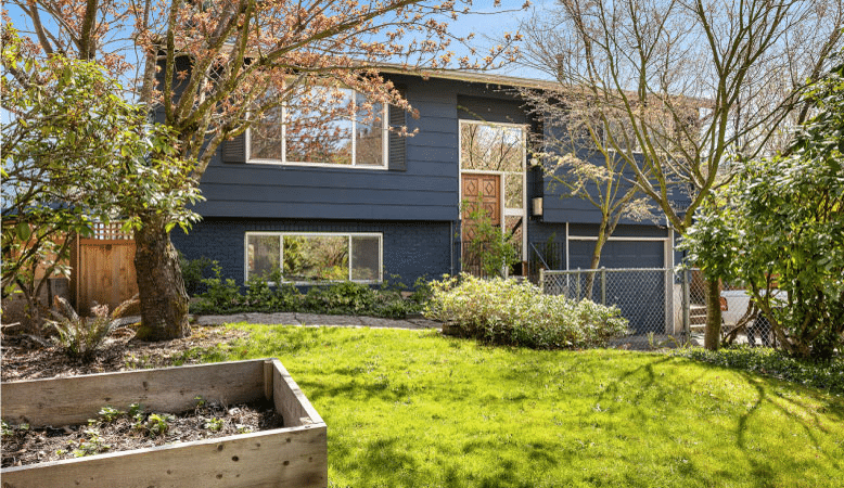 A boxy, two-story home in the St. Johns neighborhood of Portland, Oregon, The exterior is brick for the lower level and wood siding for the upper level. The entire home is painted a deep blue. There’s a raised wooden garden bed in the front yard and a variety of trees and shrubs.