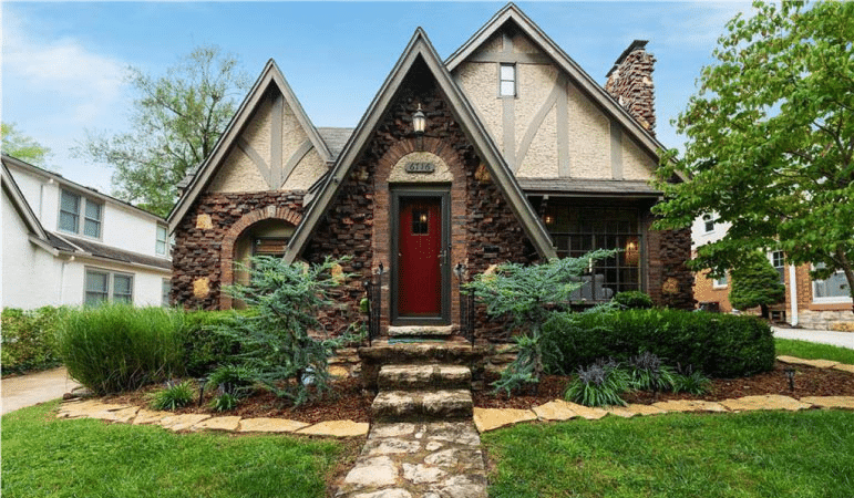 A Tudor-style cottage with beautiful landscaping in the Armour Hills neighborhood of Kansas City, Missouri. The exterior of the home is made using a stacked brick design, the door is painted red, and the walkway and stairs leading up to the home are made of natural stone pavers. 