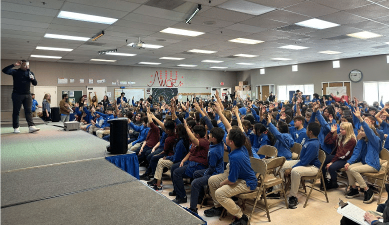 A classroom at Coral Academy of Science in Las Vegas is filled with enthusiastic students in blue and burgundy school attire. A speaker is crossing the stage holding a microphone and the students are raising their hands in response to something he said.