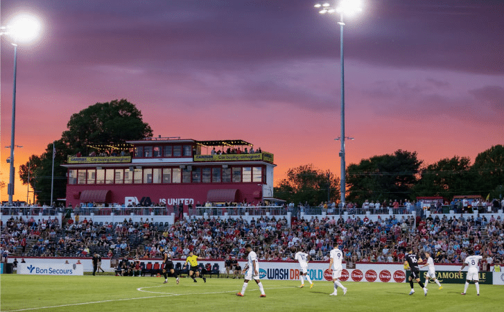 The Richmond Kickers are playing a game at City Stadium in Richmond, Virginia, in front of a full stadium. The sun is setting in the distance, turning the sky a myriad of colors from orange to dark blue.