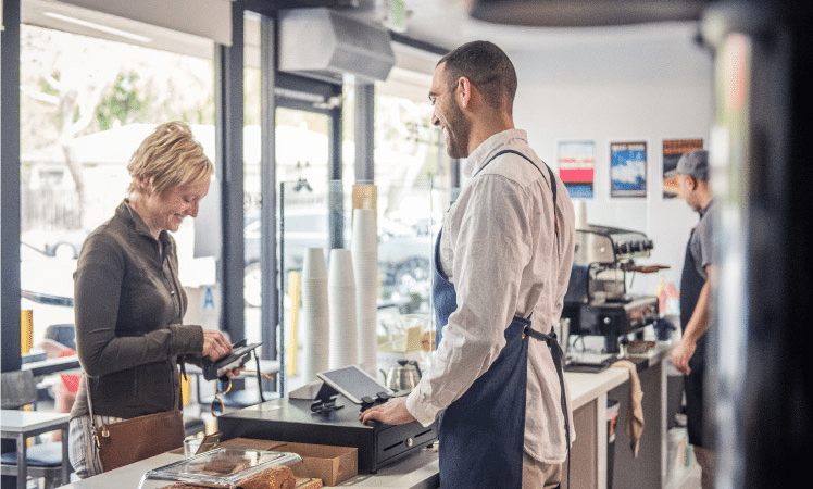 A woman is smiling as she takes money out of her wallet to pay for something at a cafe counter. The man behind the counter is wearing an apron and smiling back at the woman. Natural light is pouring into the shop through the large glass windows, and a barista can be seen standing beside an espresso machine in the back of the shop.