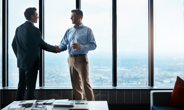 A man in a suit is shaking hands with a man in a button-up shirt and slacks, whom he just hired for a new position at the company. They’re standing in a corporate office in a tall building. Behind the men are expansive glass windows showing the surrounding area below. 