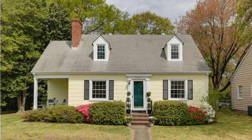 A charming Cape Cod home in the Lakeside neighborhood of Richmond, Virginia. The home is a light yellow with a dark green door and dark shutters. The yard is simple with short grass and a few bushy shrubs on either side of the entrance. 