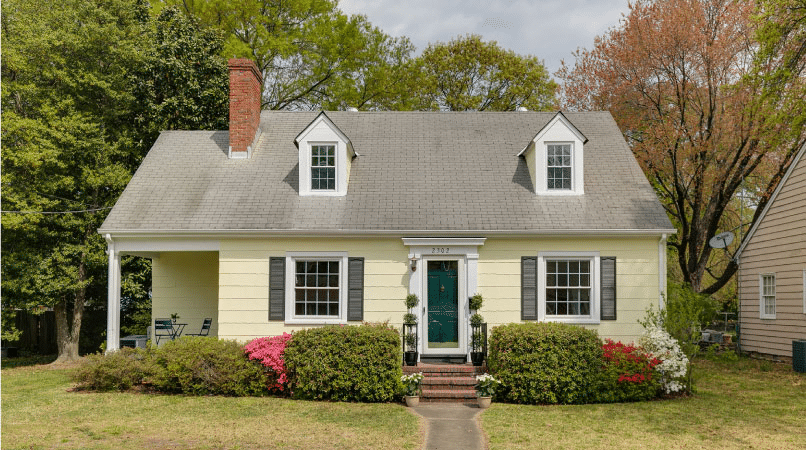 A charming Cape Cod home in the Lakeside neighborhood of Richmond, Virginia. The home is a light yellow with a dark green door and dark shutters. The yard is simple with short grass and a few bushy shrubs on either side of the entrance.