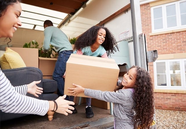 A family moving boxes out of a rental truck. A young girl hands a cardboard box to her sister.