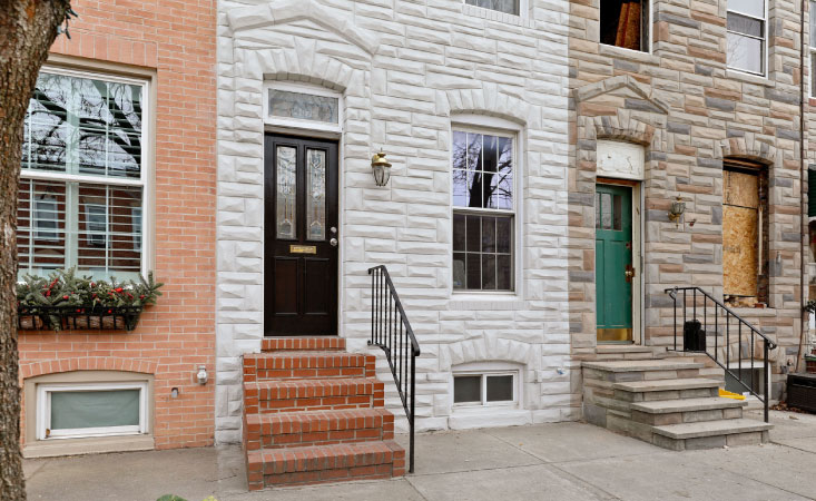 A row of homes in the Locust Point neighborhood of Baltimore, Maryland. One home is made of red brick, the second is made of painted white stone, and the third is made of multi-colored stones. 