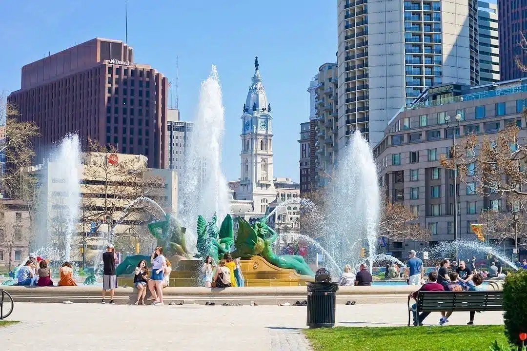 Locals are gathered around an iconic fountain in downtown Philadelphia on a sunny day — a welcome sight for those moving to Philadelphia.